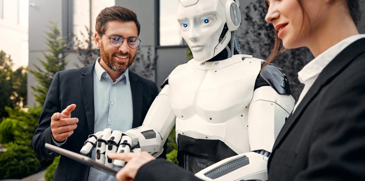 Young man and woman business partners in suits with a tablet standing together with a robot and discussing work against the background of a building. The future with artificial intelligence.