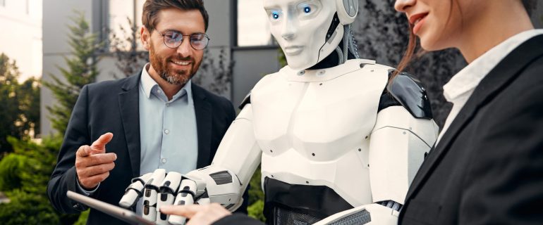 Young man and woman business partners in suits with a tablet standing together with a robot and discussing work against the background of a building. The future with artificial intelligence.