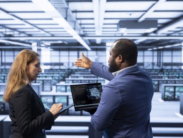 Team of technicians in data center using laptop to visualize artificial intelligence neural networks made up of interconnected nodes. Teamworking colleagues oversee AI systems processing information